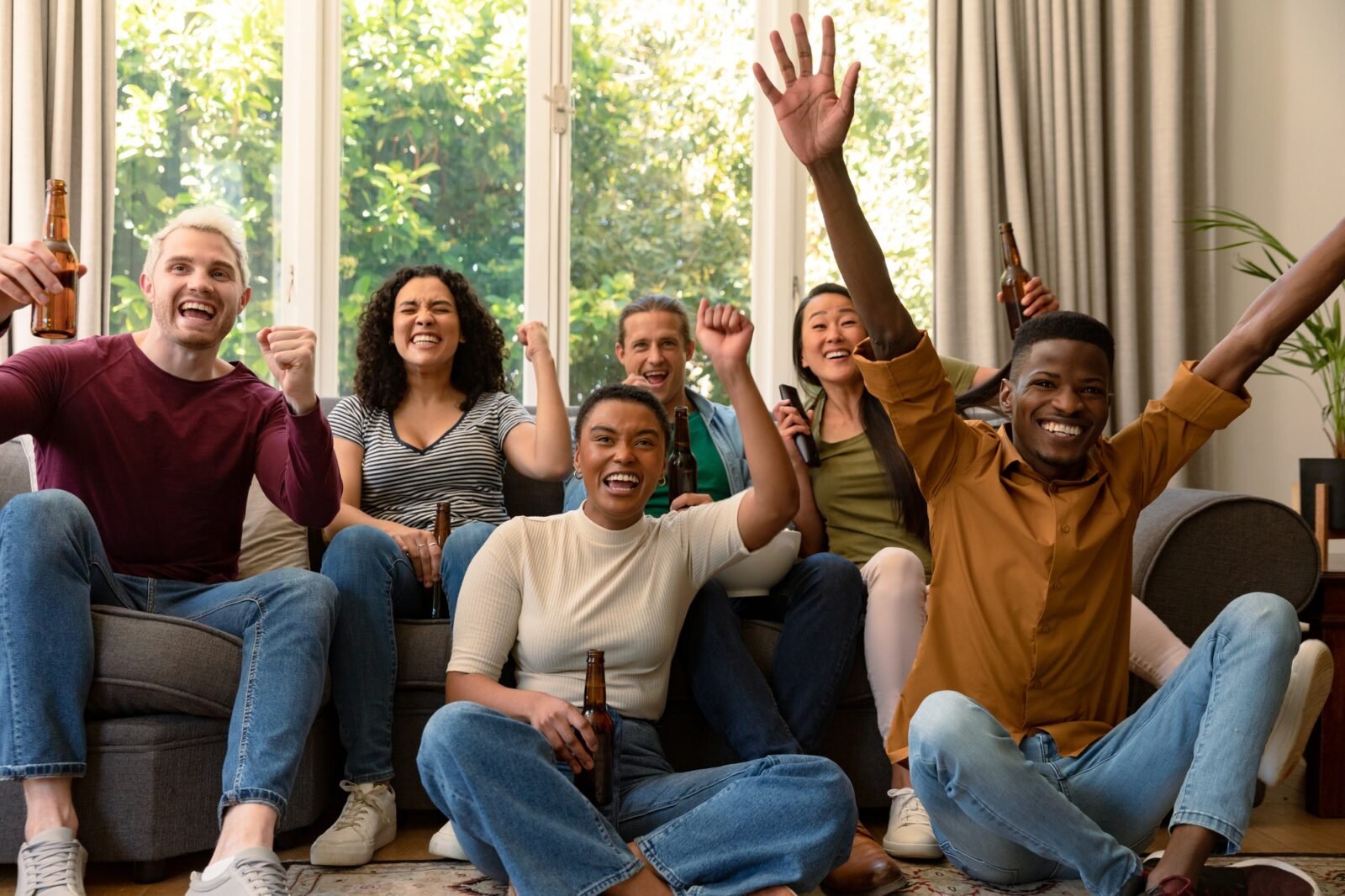 Group of happy diverse female and male friends watching tv and drinking beer together at home